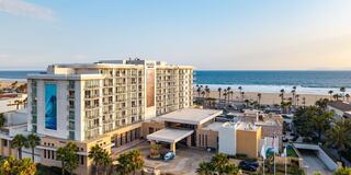 A modern hotel overlooks a sandy beach, with palm trees and ocean views, creating a scenic and inviting atmosphere.