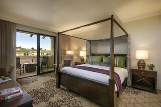 A stylish hotel room features a four-poster bed, elegant furnishings, and a balcony with a view, creating a serene and inviting atmosphere.