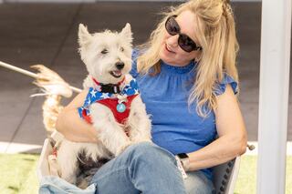 A smiling woman sits in a chair, wearing sunglasses, holding a dog in a patriotic outfit. Both seem to enjoy a sunny day together.
