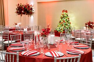 An elegantly decorated venue features red and white tables, crystal glassware, floral arrangements, and a beautifully lit Christmas tree.