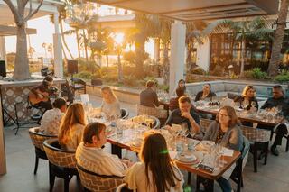 A lively restaurant scene at sunset, featuring guests enjoying meals, live music, and a warm, inviting atmosphere surrounded by palm trees.
