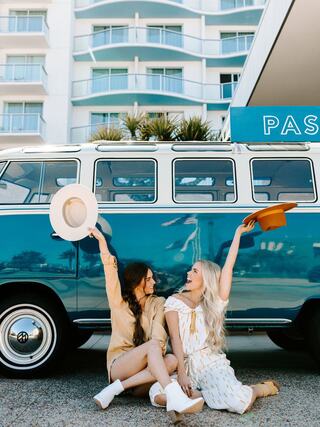Two fashionable women sit playfully in front of a vintage blue van, each holding stylish hats, exuding fun and summer vibes.