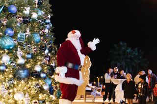 Santa waves cheerfully near a festive Christmas tree, surrounded by excited children and families enjoying the holiday spirit at night.