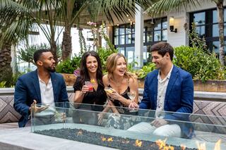 Four friends enjoy drinks by a fire pit, surrounded by greenery. Laughter and good vibes are in the air, creating a lively atmosphere.