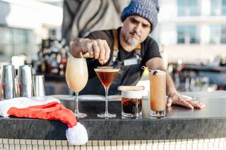 A bartender creates cocktails, featuring a festive atmosphere with a Santa hat on the bar, showcasing artistry and vibrant drinks.