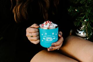 A person holds a teal mug topped with whipped cream and festive sprinkles, surrounded by holiday decorations and cozy attire.