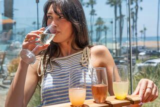 A woman enjoys a drink while sitting at a beachside venue, surrounded by light and ocean views, with a flight of colorful beverages before her.