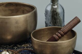 Two bronze bowls of varying sizes are displayed alongside a wooden stick and a decorative bottle, resting on a textured fabric.