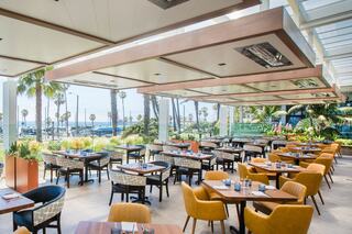 A stylish restaurant features modern furniture, ample natural light, and a view of palm trees and the ocean, creating a vibrant dining atmosphere.