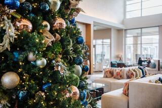 A festive Christmas tree adorned with blue and gold ornaments stands in a stylish, modern lobby with cozy seating and large windows.