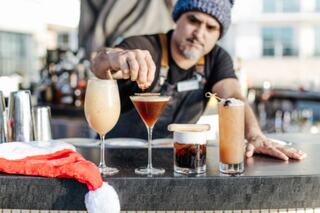 A bartender prepares festive cocktails, showcasing various drinks lined up on the bar, with a holiday-themed Santa hat nearby.
