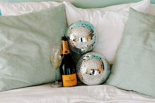 A bottle of champagne rests between two mirrored disco balls, nestled among soft, light green pillows on a cozy bed.