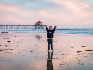 A joyful child stands in shallow water at the beach, with arms raised, reflecting colorful skies and a pier in the background.