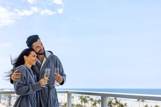 A happy couple in matching robes enjoys champagne together on a balcony overlooking a beach, radiating love and relaxation.
