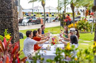 A cheerful group of friends enjoys a festive outdoor meal, raising glasses in a lush garden by a pool, surrounded by vibrant flowers.