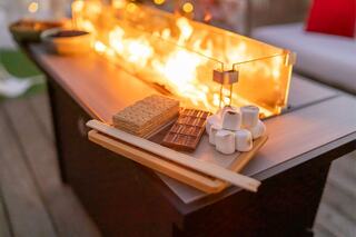 A cozy setup features chocolate bars, graham crackers, and marshmallows on a table beside a glowing fire, perfect for s'mores.