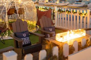 Cozy outdoor seating area features rustic chairs by a glowing fire pit, surrounded by twinkling lights and festive decorations.