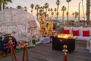 A cozy outdoor seating area features a fire pit, decorated trees, and an igloo structure, all set against a sunset by the beach.