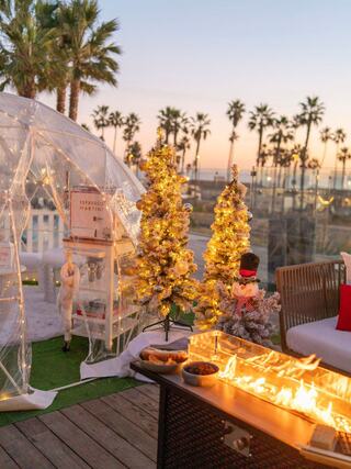 A cozy holiday scene features illuminated trees, a clear dome structure, palm trees, and a fire pit against a sunset backdrop.