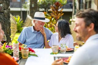 A joyful outdoor gathering features a smiling elderly couple sharing a tender moment, surrounded by friends and vibrant tropical decor.