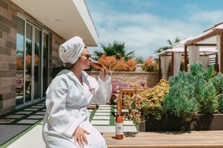A relaxed woman in a robe sips champagne outdoors, enjoying a serene spa-like atmosphere with lush greenery and sunny skies.