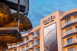 A modern hotel features a large sign reading "PASÈA Hotel & Spa," with balconies and a backdrop of a beautiful sky. Elegant lighting hangs nearby.