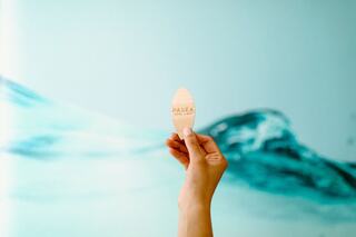 A hand holds a wooden paddle with "PASEA" engraved on it against a soft blue wall with wave-like designs.