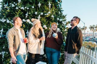 Four friends laugh and enjoy drinks in front of a festive Christmas tree, surrounded by palm trees and a sunny, coastal backdrop.