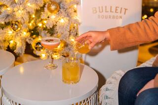 A hand pours a drink into a glass on a table beside festive cocktails, with a beautifully decorated tree glowing in the background.