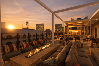 A stylish rooftop lounge features cozy seating and a fire pit, with stunning city views at sunset, creating a warm, inviting atmosphere.