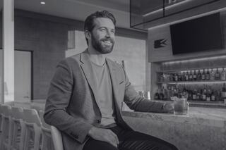 A smiling man in a stylish blazer sits at a bar, exuding confidence and charm in a modern, elegant setting.