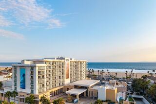 A modern hotel overlooks a sandy beach and ocean, surrounded by palm trees under a clear blue sky. Perfect for a coastal getaway.
