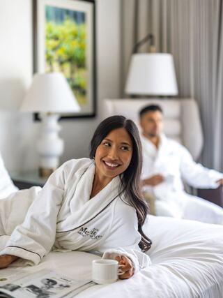 A woman in a cozy robe smiles while lounging on a bed, holding a cup, as a man relaxes in the background. Light and comfort set the scene.