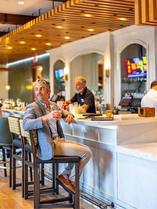 A man in a suit casually sits at a bar, enjoying a drink while bartenders prepare beverages in a stylish, modern setting.