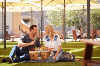A couple enjoys a picnic on a grassy lawn, sharing laughter and drinks under large umbrellas, creating a warm, intimate atmosphere.