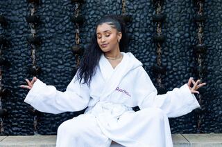A woman in a white robe sits in a tranquil pose, eyes closed, in front of a decorative water feature, exuding calmness and relaxation.
