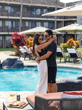 A couple embraces by a pool, surrounded by vibrant flowers and lounge chairs, enjoying a sunny day and each other's company.
