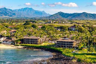 A scenic coastal view features lush palm trees and beachfront buildings, with majestic mountains rising in the background under a clear sky.