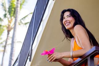 A smiling woman with long hair holds a pink flower while leaning on a balcony, surrounded by palm trees under a clear blue sky.
