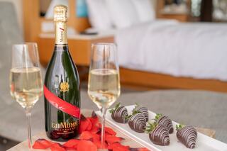A bottle of champagne sits between two flutes, accompanied by chocolate-covered strawberries and rose petals, creating a romantic setting.