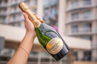 A hand holds a bottle of Perrier-Jouët champagne, showcasing its elegant label against a blurred modern building background.