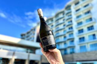 A hand holds a bottle of sparkling wine against a sunny backdrop of a modern building, conveying a festive and celebratory atmosphere.