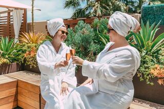 Two women in white robes and towels celebrate with champagne glasses amidst lush greenery in a sunny, relaxed setting.