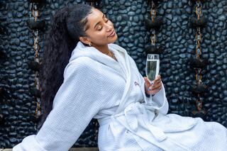 A woman in a cozy white robe relaxes with a glass of sparkling beverage, exuding a sense of luxury and tranquility against a textured backdrop.
