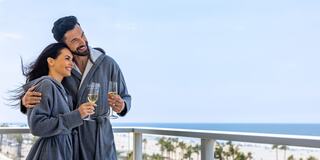 A couple in cozy robes enjoys champagne on a balcony, smiling and embracing, with a beach view in the background.