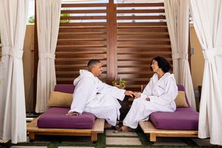 A couple in matching robes sits on lounge chairs, smiling and holding hands, surrounded by a cozy, serene outdoor setting.