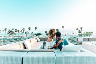 A couple embraces on a stylish outdoor sofa, surrounded by palm trees and a serene coastal view, exuding romance and tranquility.