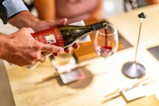 A hand pours red wine from a bottle into a glass, while hints of a dining setting create an elegant atmosphere.