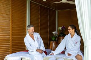 A man and woman in white robes share a relaxed moment in a serene spa setting with wooden decor and tropical plants around them.