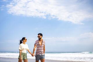 A couple walks hand in hand along a sunny beach, enjoying each other's company against a backdrop of waves and a clear sky.